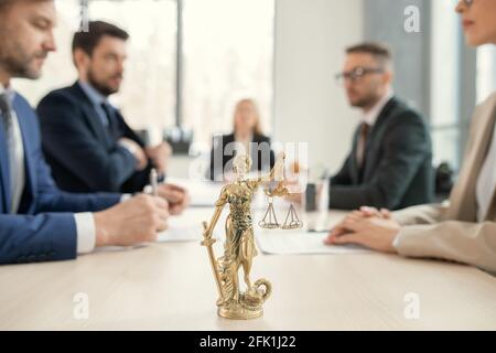 Konzentrieren Sie sich auf Golden Themis mit Gerechtigkeitsskalen und Sward auf dem Tisch der Anwälte, Kollegen sitzen einander im Hintergrund gegenüber Stockfoto