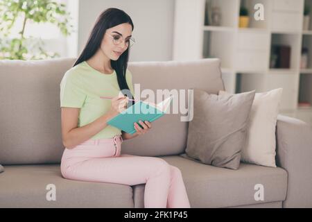 Foto von fokussierten Dame sitzen Sofa halten Lehrbuch Stift Notizblock Schreiben Tragen Sie Brillen grün T-Shirt rosa Hosen drinnen Stockfoto