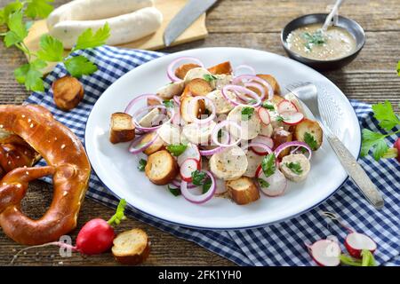 Herzhafter bayerischer Salat mit geschnittenen Kalbswürsten, gebratenen Brezelscheiben und einem Senfdressing auf einem rustikalen Holztisch Stockfoto