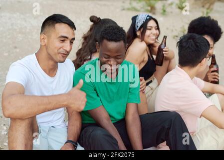 Eine Gruppe von fröhlichen Freunden, die in Sommerkleidung gekleidet sind und am Strand mit Bierflaschen rösten. Stockfoto