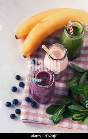 Drei bunte Smoothies in Flaschen mit Strohhalm auf weißem Tisch. Banane, Blaubeeren und grüne Minzblätter. Gesundes veganes Essen. Draufsicht. Stockfoto