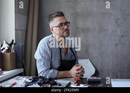 Linolschnitt-Meister in Schürze oder Handwerker, der am Arbeitsplatz sitzt und über ein neues Projekt nachdenkt. Bärtiger männlicher Künstler mittleren Alters in einer Brille, die auf einem Hintergrund des Atelierarbeitsplatzes sitzt. Hochwertige Fotos Stockfoto