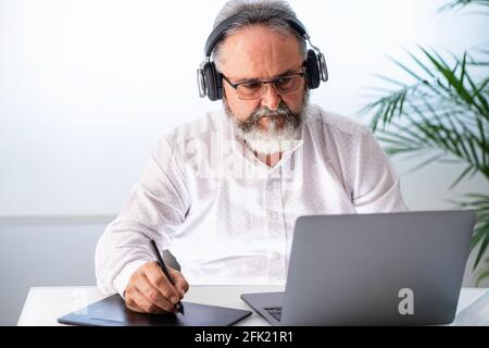 Ein älterer Mann mit Kopfhörern, der mit einem Laptop und einem Tablet arbeitet Zu Hause Stockfoto