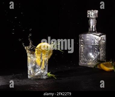 Farbloses alkoholisches Getränk in einem Glas mit einer Scheibe Zitrone und Rosmarin, Spritzer. Wodka und Dekanter auf schwarzem Hintergrund. Stockfoto