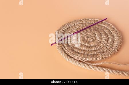 Spiralförmige Jute-Serviette und Häkelnadel auf beigem Hintergrund. Hobbys, Handarbeiten. Stockfoto