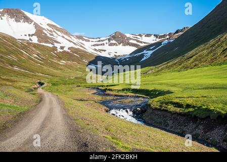 Verlassene, kurvenreiche, raue Straße entlang eines Baches am Fuß Von schneebedeckten Gipfeln in einer majestätischen Berglandschaft auf einem Sonniger Sommertag Stockfoto