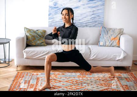 Glückliche afroamerikanische junge Frau in sportlicher schwarzer Kleidung beim Aufwärmen im Wohnzimmer, stürzt sich vorwärts, die Handflächen sind gefaltet, macht Fitness, führt einen gesunden Lebensstil, schaut auf die Kamera, lächelt Stockfoto