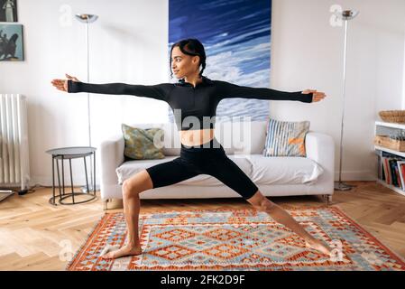 Aktive junge afroamerikanische Frau mit sportlicher Figur, in Sportbekleidung, macht ein Warm-up, steht im Wohnzimmer, macht zu Hause Fitness, führt einen gesunden Lebensstil, schaut lächelnd zur Seite Stockfoto