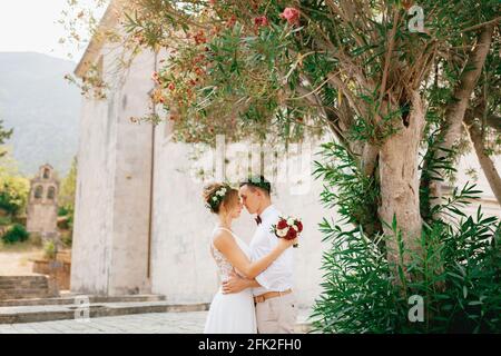 Die Braut und der Bräutigam umarmen sich zärtlich in der Nähe des blühenden Oleanderbusches in der Nähe der alten Kirche in Prcanj und halten einen Strauß in der Hand Stockfoto