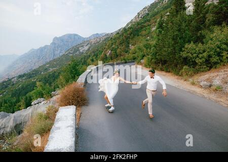 Die Braut rollt auf einem Skateboard und hält die Hand des Bräutigams, der entlang der Autobahn in den Bergen läuft Stockfoto