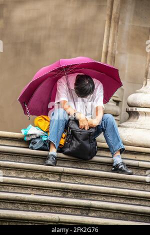 Tourist mit Sonnenschirm - Mann sitzt auf der Treppe neben Riesige Säule über Rucksack unter rosa Regenschirm gebeugt - müde Oder gedrückt oder gelangweilt oder überprüft Stockfoto