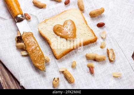 Hausgemachtes Toast zum Frühstück mit frischer, knuspriger, herzförmiger Erdnussbutter und Nüssen auf hellem Textilhintergrund. Stockfoto