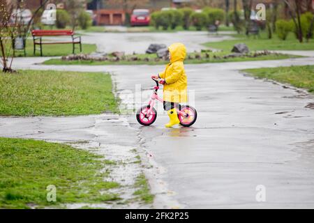 Kleines Mädchen in gelber Regenjacke fährt bei Regen auf einer Fahrradtour durch Pfützen. Regnerisches Wetter im Frühling. Kind lernt, den zweirädrigen Transport zu bewältigen. Lear Stockfoto