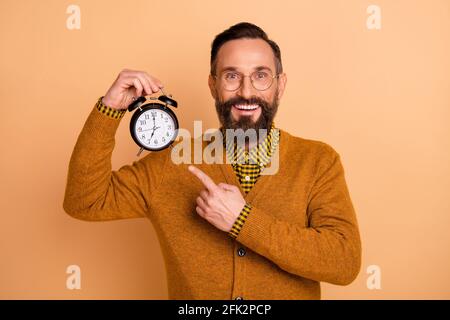 Foto von optimistischen braunen Haaren Mann Point Clock tragen Brille Brauner Pullover isoliert auf beigefarbenem Hintergrund Stockfoto