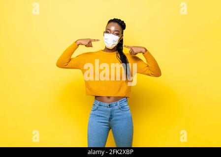 Junge hygienische gesunde afrikanische Frau trägt und zeigt ins Gesicht Maske im gelben, isolierten Studiohintergrund Stockfoto