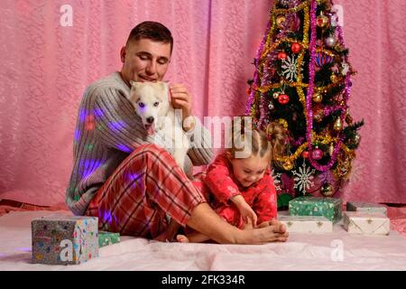 Papa mit Tochter und Husky Welpe sitzt am Weihnachtsbaum, Papa und Tochter im Pyjama. Neu Stockfoto