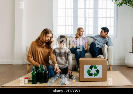 Kaukasischer Bruder und Schwester setzen Kunststoff- und Glasflaschen ein Pappkarton zum Recycling, während Mutter und Vater sitzen Couch und reden zu Hause Stockfoto