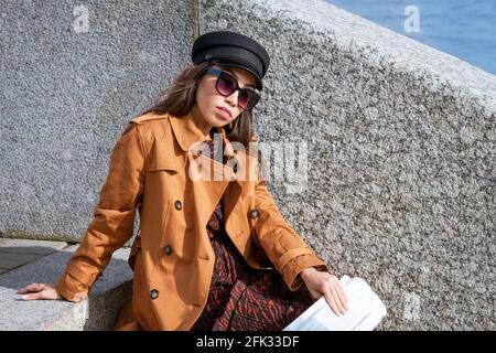 Junge Frau in Sonnenbrille der kaukasischen Ethnie sitzt auf dem Schritte auf dem Damm in einer schwarzen Kappe und braun Jacke mit Zeitung in der Hand Stockfoto