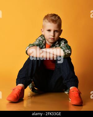 Blonde Teenager, normaler Kerl im karierten Hemd, orangefarbenes T-Shirt und Jeans sitzen auf dem Boden und schauen auf die Kamera Stockfoto