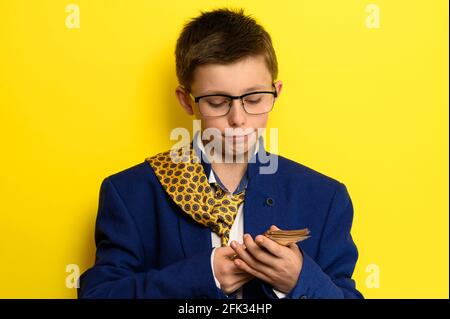 Ein 11-jähriger Junge in einem übergroßen Anzug hält einen Euro, ein Porträt eines Kindes mit Geld auf gelbem Hintergrund. Stockfoto