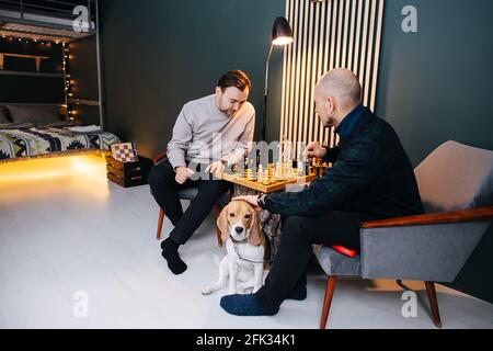 Männer mittleren Alters, die zu Hause Schach spielen. Einer von ihnen spielt mit einem Hund. Neben einem Stumptisch unter einer Lampe sitzend. Stockfoto