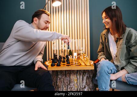 Lächelndes Paar mittleren Alters, das zu Hause ein Schachspiel spielt und Spaß hat. Sitzen neben einem Stumptisch unter einer Lampe in Komfort von zu Hause. Stockfoto