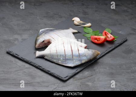 Roher weißer Pomfrett, fertig zum Kochen Pomfrett auf einem Rechteck angeordnet Form Graphitblech mit Tomaten, Blätter und Knoblauch wie Einige Zutaten zum Kochen Stockfoto