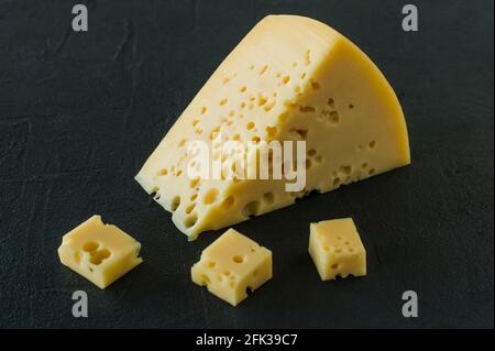Radamer-Käse auf schwarzem Betongrund. Dreieckiges Stück gelber Schweizer Kuhmilch-Käse mit Löchern. Stockfoto