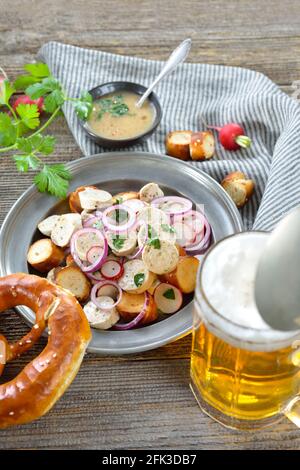 Herzhafter bayerischer Salat mit geschnittenen Kalbswürsten, gerösteten Brezelscheiben auf einem Blechteller, serviert mit einem Senfdressing und einem Becher Lagerbier Stockfoto