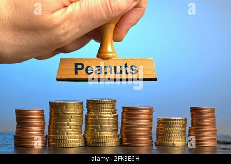 Deutschland. April 2021. Eine Holzmarke mit der Aufschrift 'Peanuts', die von einer Hand im Schnitt über mehrere Stapel Euro/Cent-Münzen auf einer glänzenden Schieferplatte gehalten wird. Foto mit Komposition vor blauem Hintergrund. Erdnüsse sind umgangssprachlich ein Ausdruck für kleine Dinge oder unbedeutende Geldsummen. Das Wort wurde zum deutschen Wort des Jahres 1994 erklärt.Quelle: dpa/Alamy Live News Stockfoto