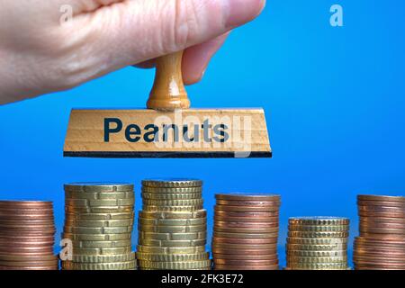 Deutschland. April 2021. Eine Holzmarke mit der Aufschrift 'Peanuts', die von einer Hand im Schnitt über mehrere Stapel Euro/Cent-Münzen gehalten wird. Foto mit Komposition vor blauem Hintergrund. Erdnüsse sind umgangssprachlich ein Ausdruck für kleine Dinge oder unbedeutende Geldsummen. Das Wort wurde zum deutschen Wort des Jahres 1994 erklärt.Quelle: dpa/Alamy Live News Stockfoto