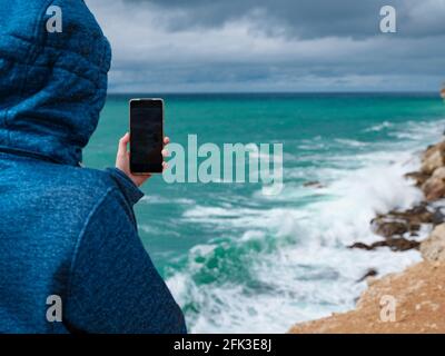 Eine Frau fotografiert das Meer auf dem Smartphone, eine Frau reist in der kalten Jahreszeit entlang der Küste Stockfoto