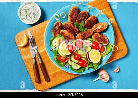 Lamm-Kofta-Kebabs aus gemahlenem Lamm, serviert auf einem blauen Teller mit Joghurtdressing, frischem Gemüsesalat und Zitronenkeilchen, griechische Küche auf einem weißen Holz Stockfoto