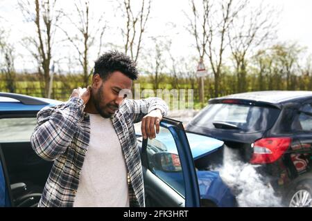 Unversicherter männlicher Fahrer, der sich bei Schmerzen durch Schleudertrauma den Hals reibt Stehen von beschädigten Auto nach Verkehrsunfall, für die er Ist schuld Stockfoto