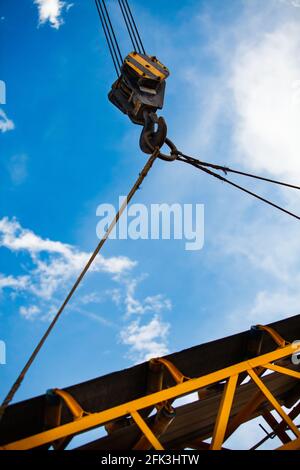 Gelber Kranhaken mit Stahlseil, der gelben Stahlträger hebt. Am blauen Himmel mit Wolken Hintergrund. Sonniger Tag. Stockfoto