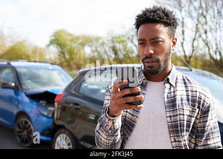 Junger Mann, der nach einem Anruf bei einem Verkehrsunfall an einem beschädigten Auto steht Panne Recovery-Unternehmen mit Mobiltelefon Stockfoto