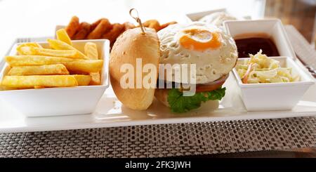 Burger mit einem Spiegelei garniert. Das Gericht wird mit Pommes Frites, einer Seite mit Soleslaw und Zwiebelringen serviert. Stockfoto