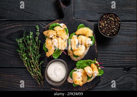 Toasts mit knusprig gebratenen Garnelen und grünem Salat auf einem Holzbrett. Schwarzer Holzhintergrund. Draufsicht Stockfoto