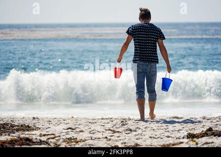 Ein junger Mann an einem sonnigen Strand, der in zwei kleinen roten und blauen Eimern Wasser für seine Sandburg holt Stockfoto