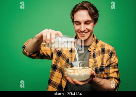 Der junge Mann gießt Milch in Flocken und sieht glücklich aus, isoliert auf grünem Hintergrund Stockfoto