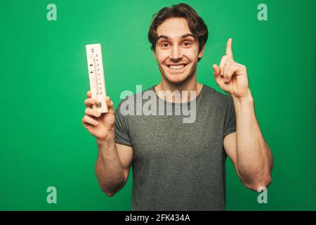 Der junge Mann erinnerte sich an etwas und hält ein Thermometer, isoliert auf grünem Hintergrund Stockfoto