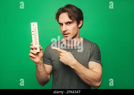 Junger Mann zeigt auf Thermometer, isoliert auf grünem Hintergrund Stockfoto