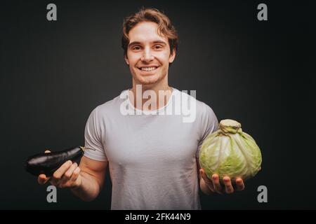 Der junge blonde Mann in weißem T-Shirt hält Auberginen und Kohl, isoliert auf schwarzem Hintergrund Stockfoto