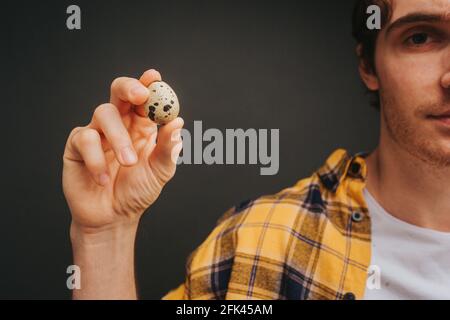 Nahaufnahme junger blonder Mann in gelbem Hemd hält Wachtelei, isoliert auf schwarzem Hintergrund Stockfoto