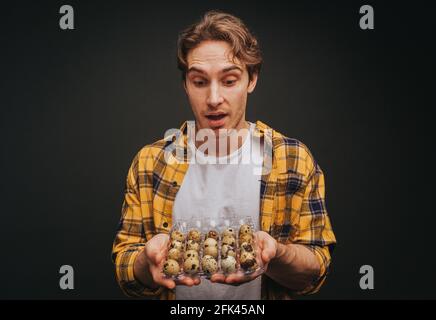 Der junge blonde Mann in gelbem Hemd hält Wachteleier, isoliert auf schwarzem Hintergrund Stockfoto