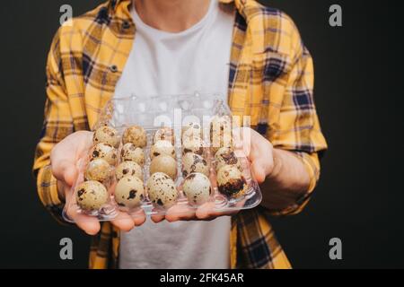 Nahaufnahme des jungen blonden Mannes in gelbem Hemd hält Wachteleier Verpackung, isoliert auf schwarzem Hintergrund Stockfoto