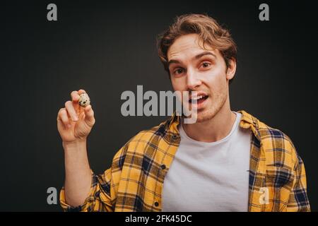 Der junge blonde Mann in gelbem Hemd hält Wachtelei, isoliert auf schwarzem Hintergrund Stockfoto