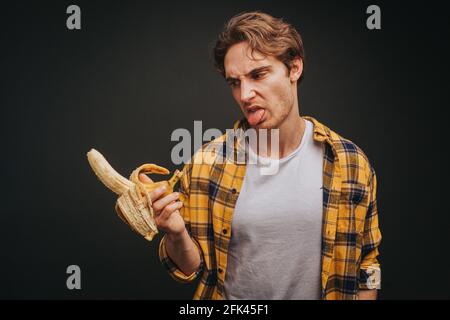Der junge blonde Mann in gelbem Hemd frisst widerwillig Banane, isoliert auf schwarzem Hintergrund Stockfoto