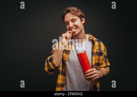 Der junge blonde Mann im gelben Hemd frisst Chips, isoliert auf schwarzem Hintergrund Stockfoto