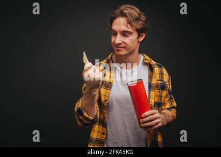 Der junge blonde Mann in gelbem Hemd hält Chips, isoliert auf schwarzem Hintergrund Stockfoto